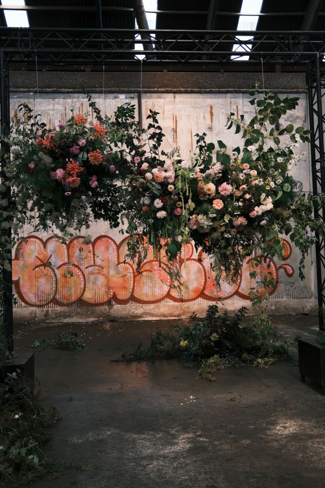 Bloemstukken hangen aan een stalen frame in een verlaten fabriekshal, voor een betonnen muur met oranje graffiti