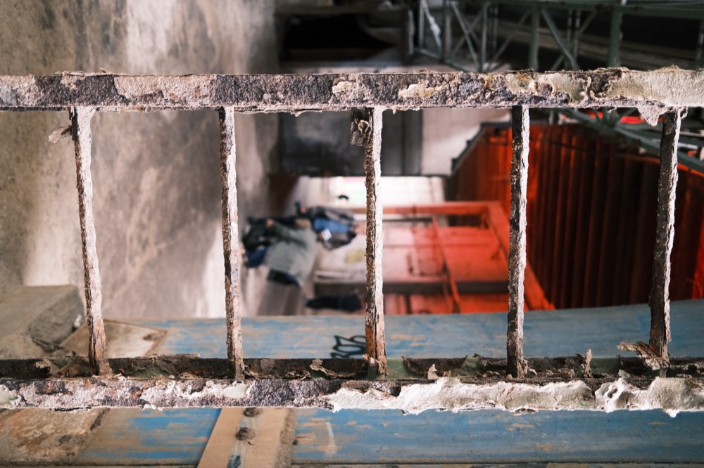 Doorkijk door een roestige ladder naar mensen in een lege fabriekshal