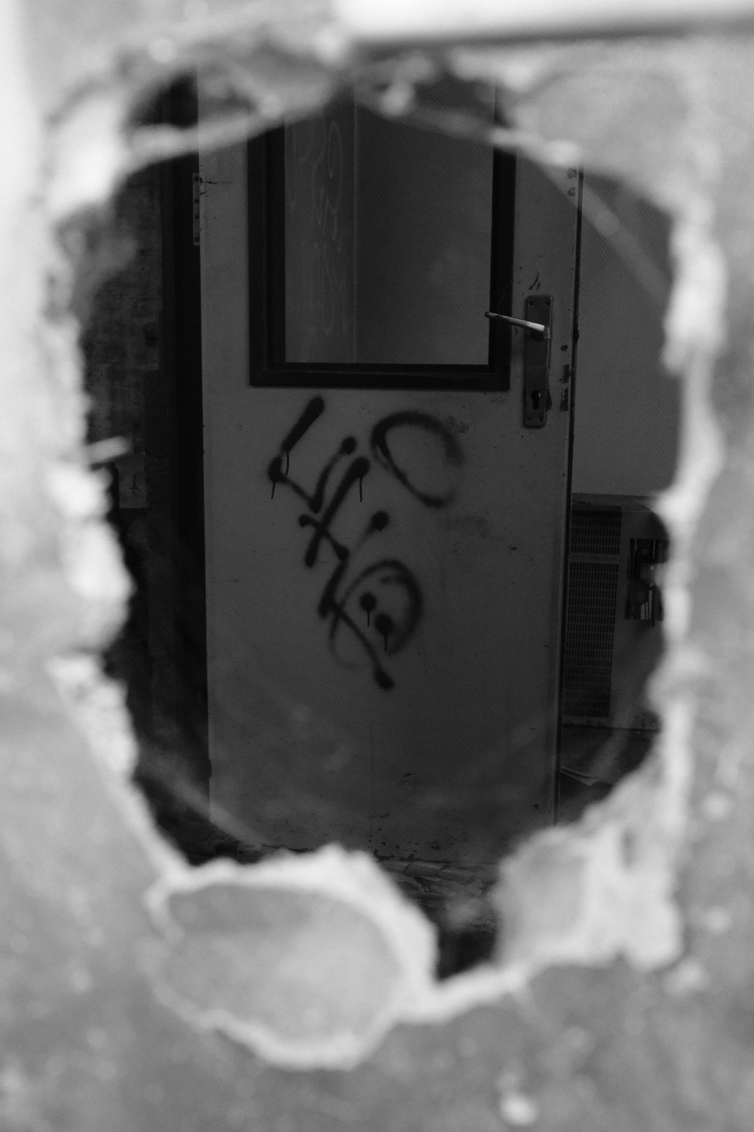 Doorkijk door een gat in een muur naar een deur met graffiti