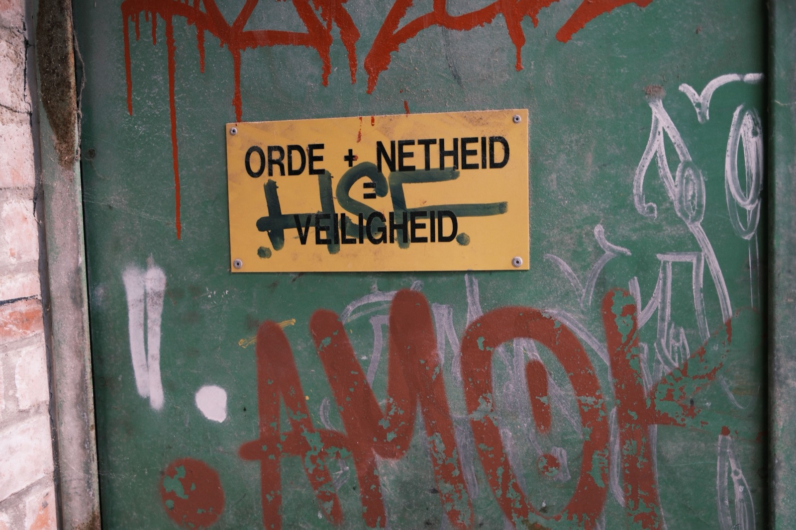 Geel bordje met de tekst 'orde + netheid = veiligheid' op een groene, met graffiti bespoten fabrieksdeur