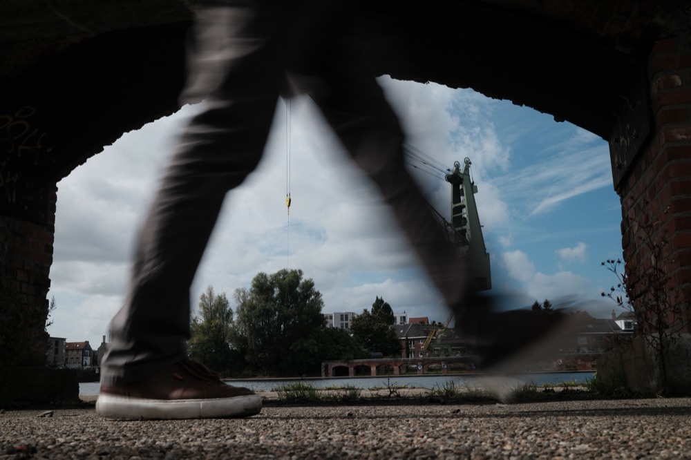 Wandelaar in beweging onder een bakstenen brug, met een oude havenkraan op de achtergrond